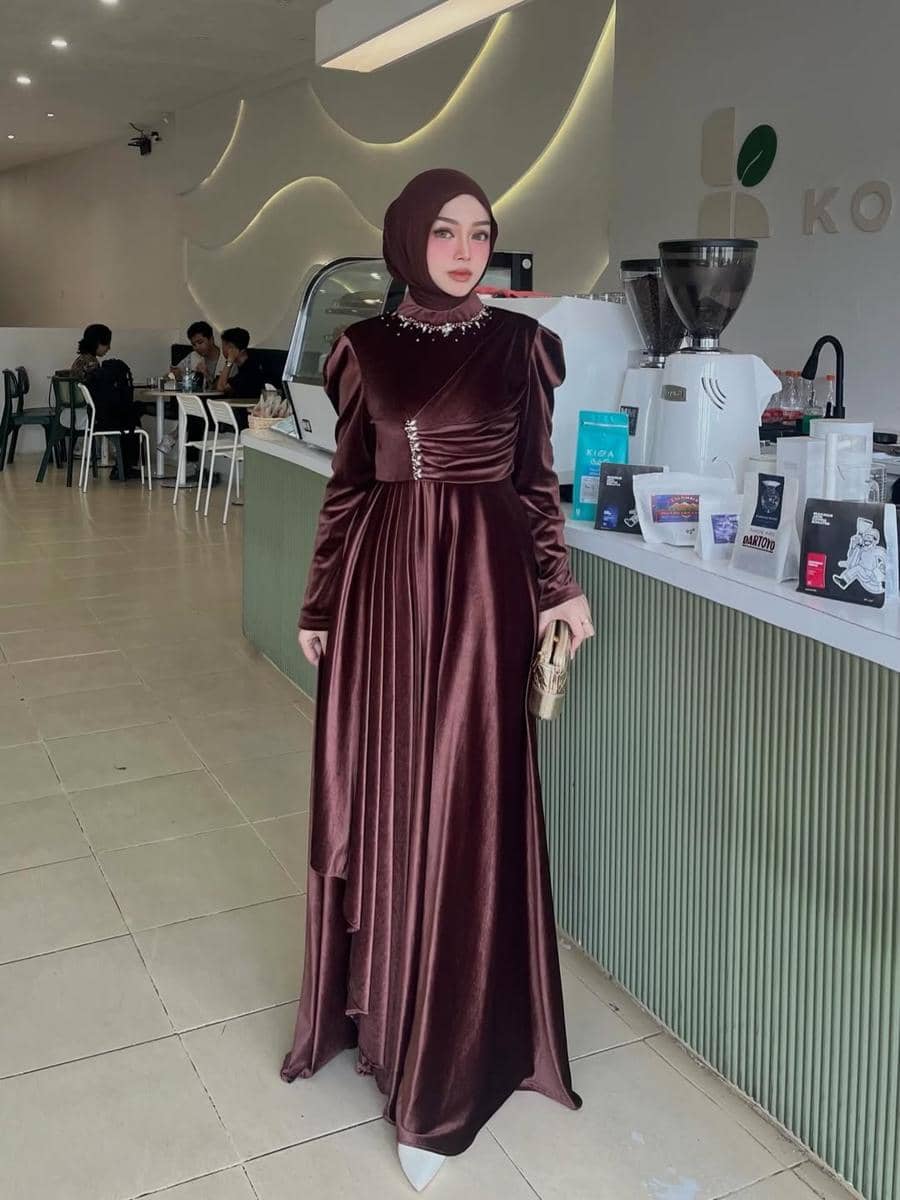 Seorang wanita berhijab mengenakan gaun panjang beludru berwarna cokelat tua dengan detail elegan berdiri di area kafe modern.