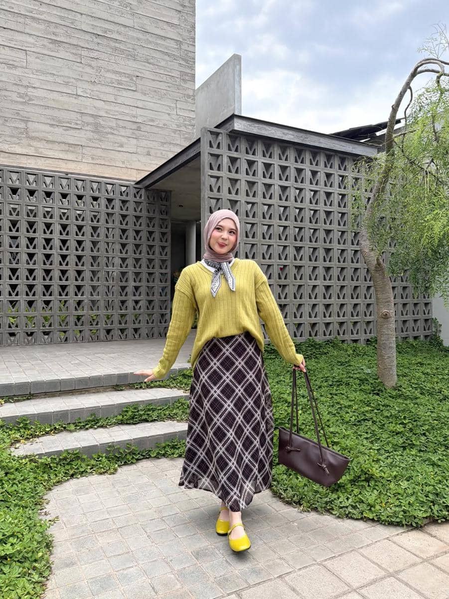 Seorang wanita berhijab mengenakan sweater hijau zaitun, rok motif kotak, sepatu kuning, dan tas cokelat berpose di depan bangunan modern.