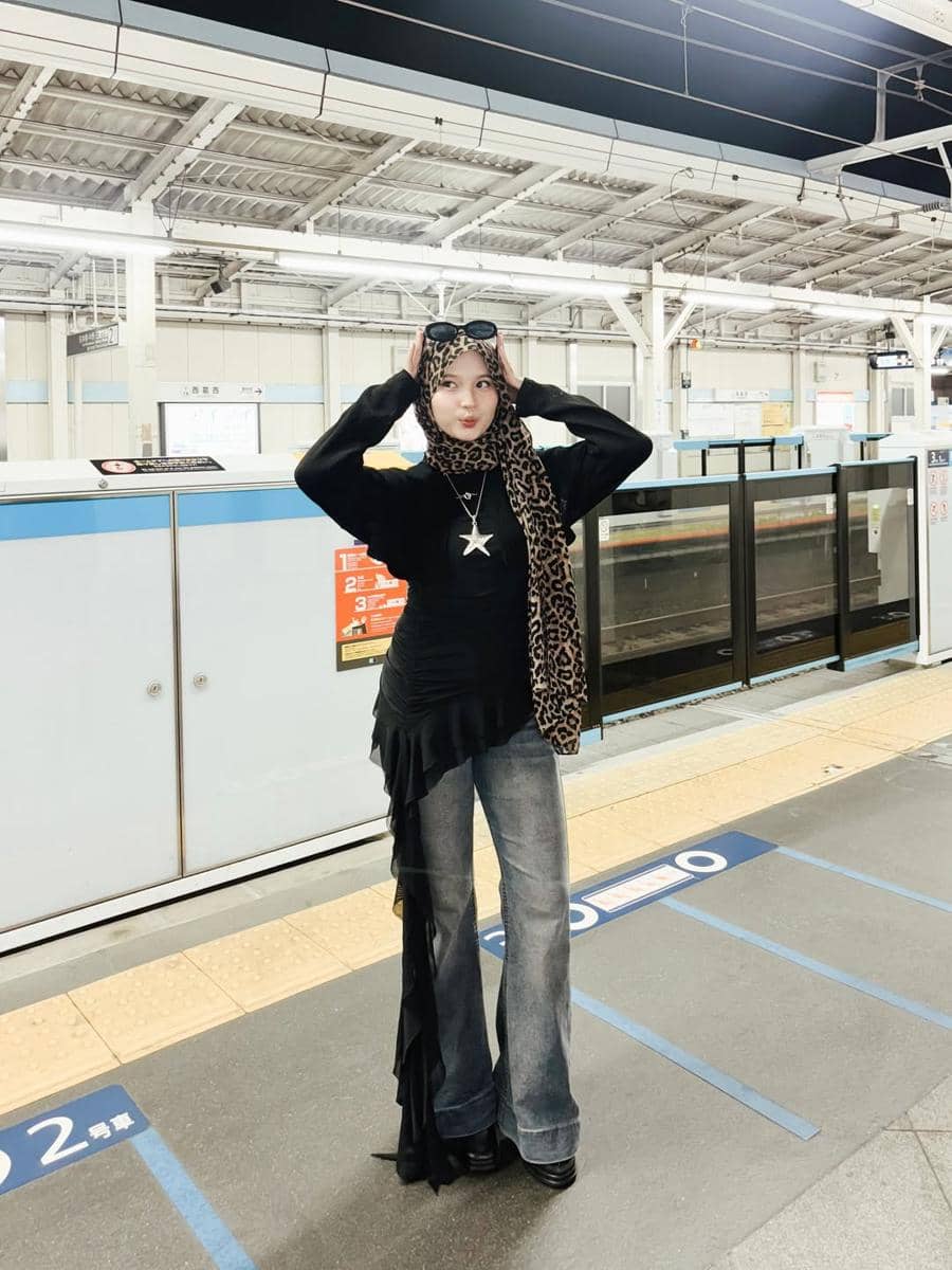 Seorang wanita berhijab bergaya modis dengan atasan hitam, celana jeans, dan syal motif leopard berpose di stasiun kereta luar negeri.