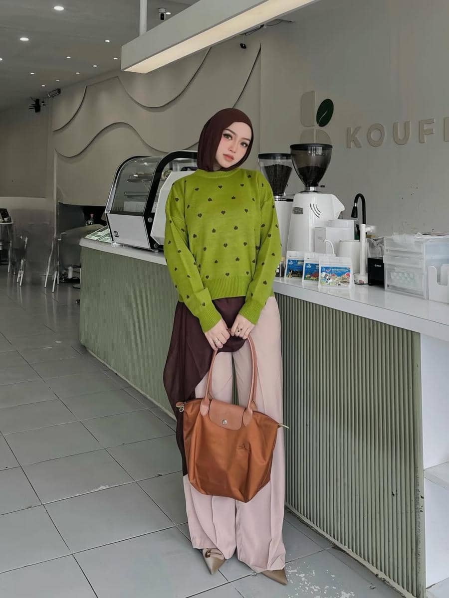Perempuan berhijab mengenakan sweater hijau bermotif dan celana krem, berpose di dalam kafe modern dengan tas cokelat besar.