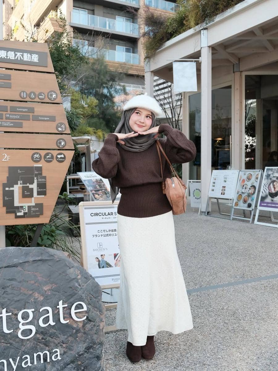 Seorang wanita berhijab mengenakan sweater cokelat, rok putih panjang, dan topi bulu putih berpose di area luar gedung modern.