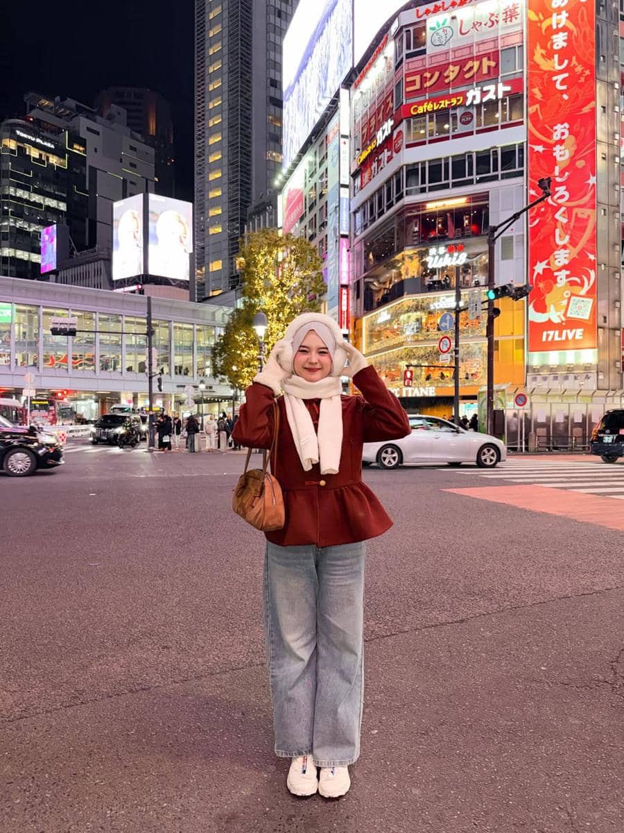 Seorang wanita berhijab mengenakan jaket merah marun dan syal putih berpose di persimpangan kota besar dengan lampu neon berwarna-warni pada malam hari.