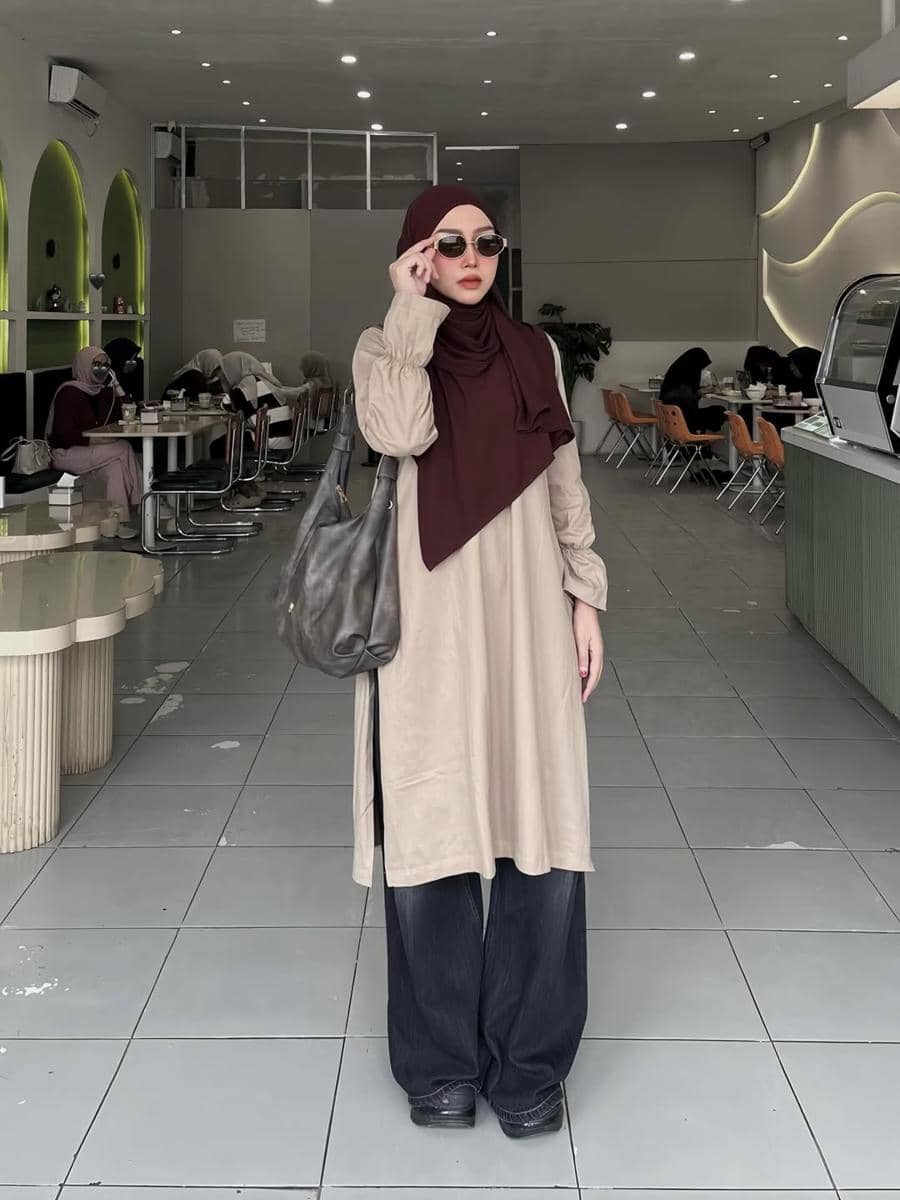 Seorang wanita berhijab marun mengenakan tunik krem dan celana hitam lebar berpose di dalam kafe modern dengan pencahayaan lembut.