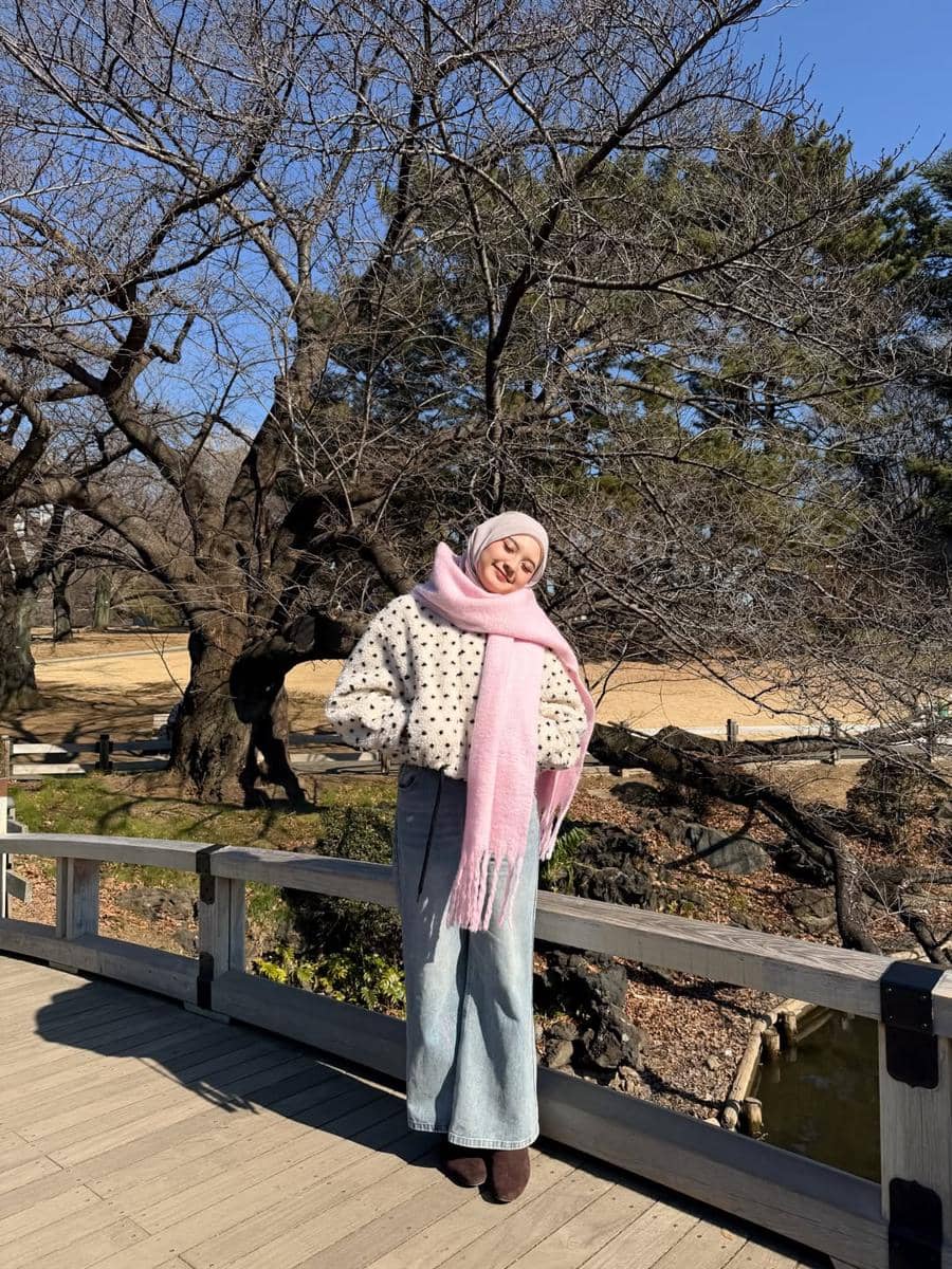 Seorang wanita berhijab mengenakan jaket tebal bermotif polkadot dan syal pink berpose di jembatan kayu saat musim dingin.