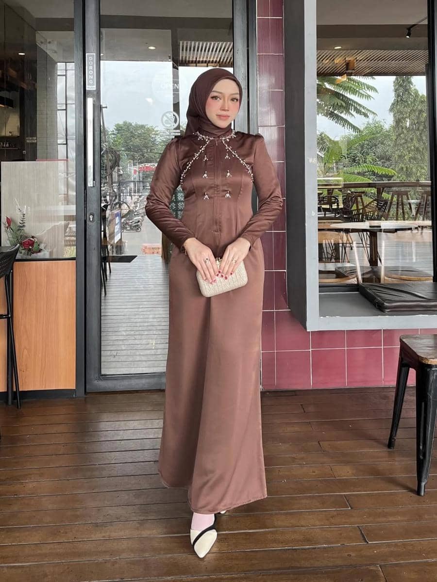 Seorang wanita berhijab mengenakan gaun panjang berwarna cokelat dengan hiasan manik di bagian dada, memegang clutch krem di depan kafe.