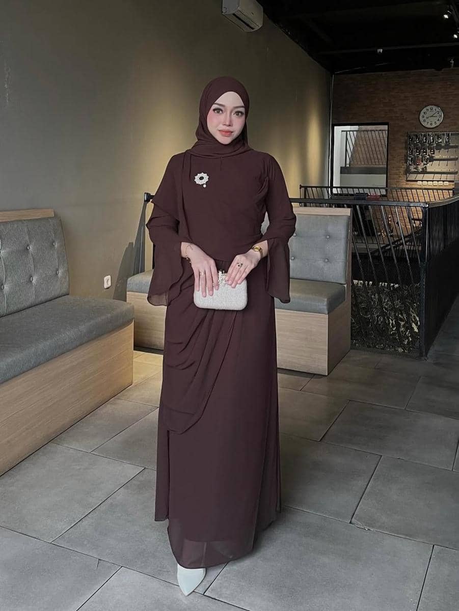 Seorang wanita berhijab mengenakan gaun panjang berwarna cokelat tua dengan tas tangan kecil berwarna krem di ruangan bergaya modern.