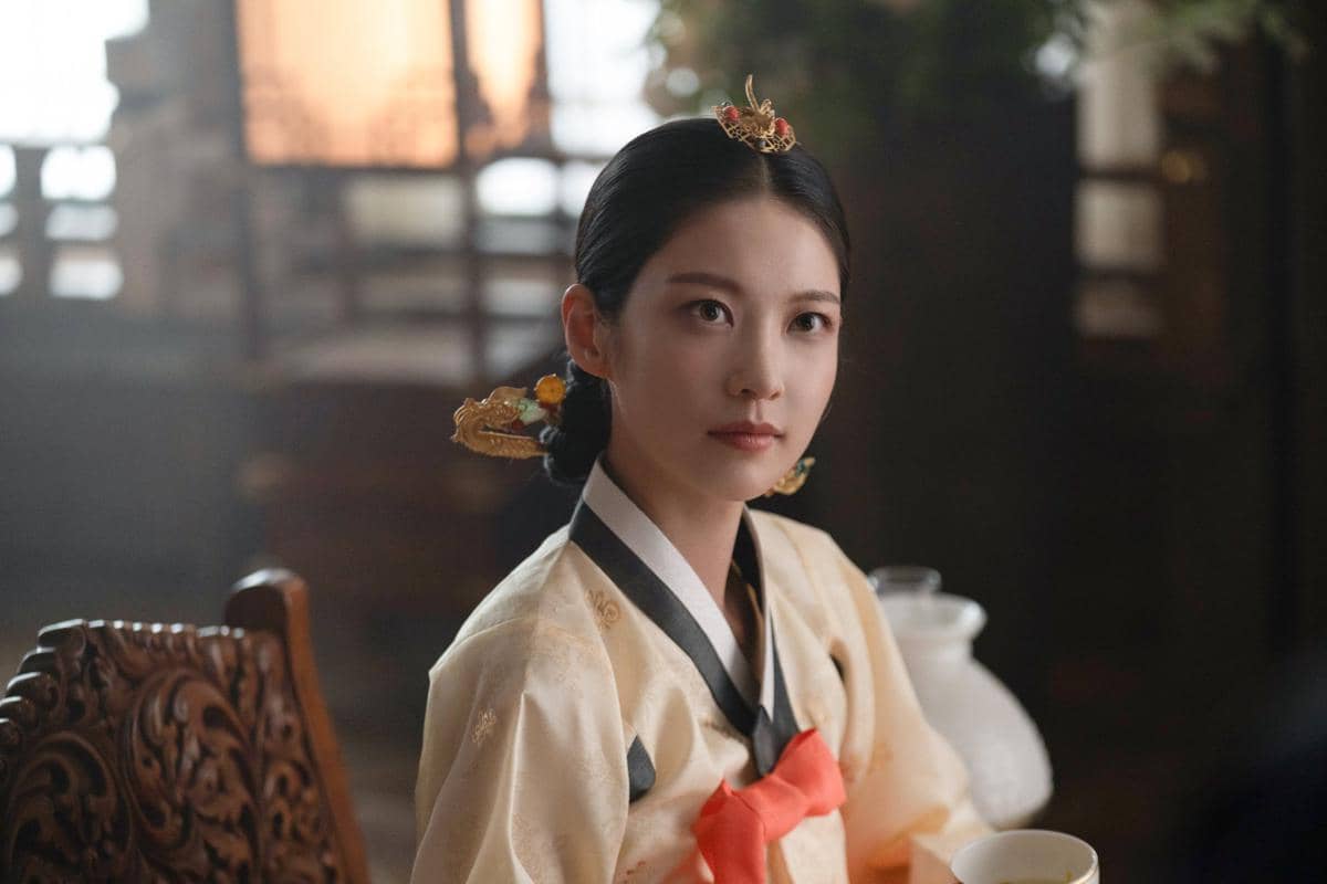 Seorang wanita mengenakan hanbok tradisional Korea berwarna krem dengan pita merah dan hiasan rambut emas duduk di dalam ruangan bergaya klasik.
