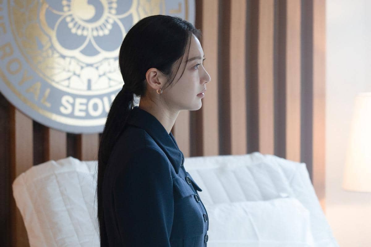 Seorang wanita berambut panjang mengenakan setelan biru gelap duduk di ruangan dengan latar logo bertuliskan Royal Seoul.