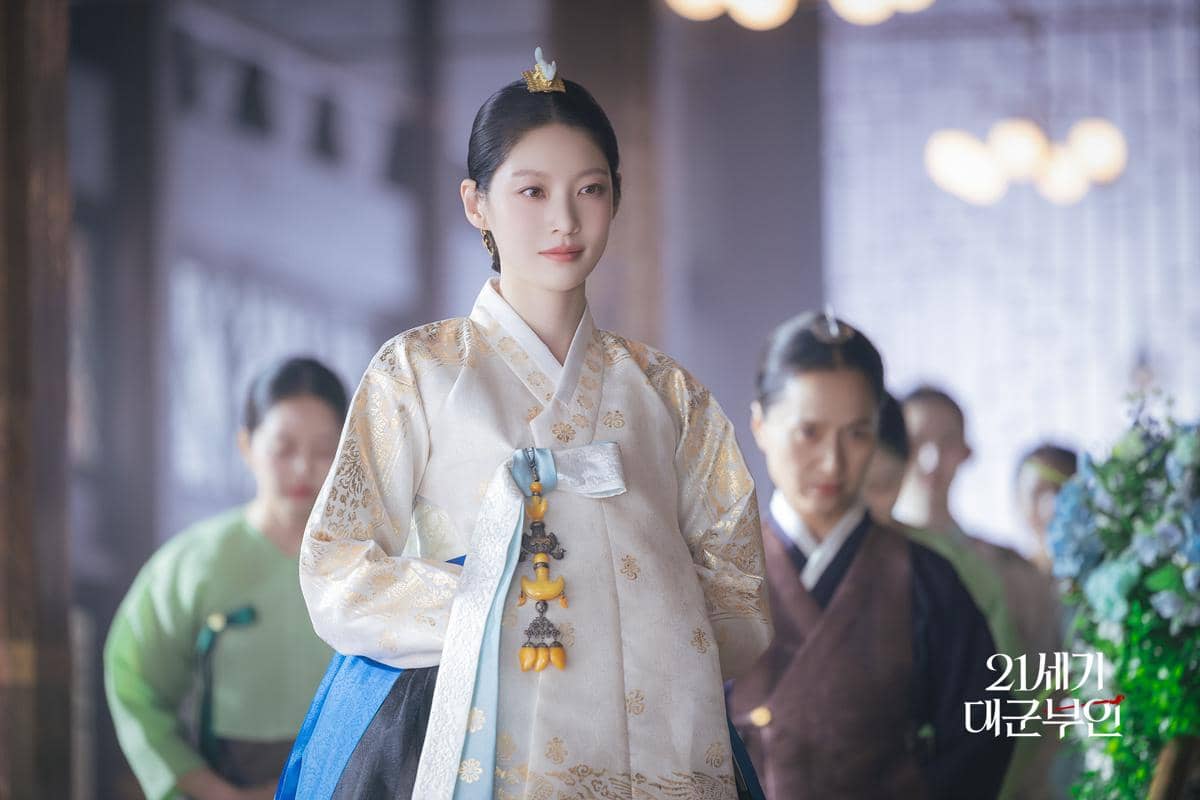 Seorang wanita mengenakan hanbok tradisional berwarna putih dan biru dengan hiasan rambut emas dalam adegan drama Perfect Crown.