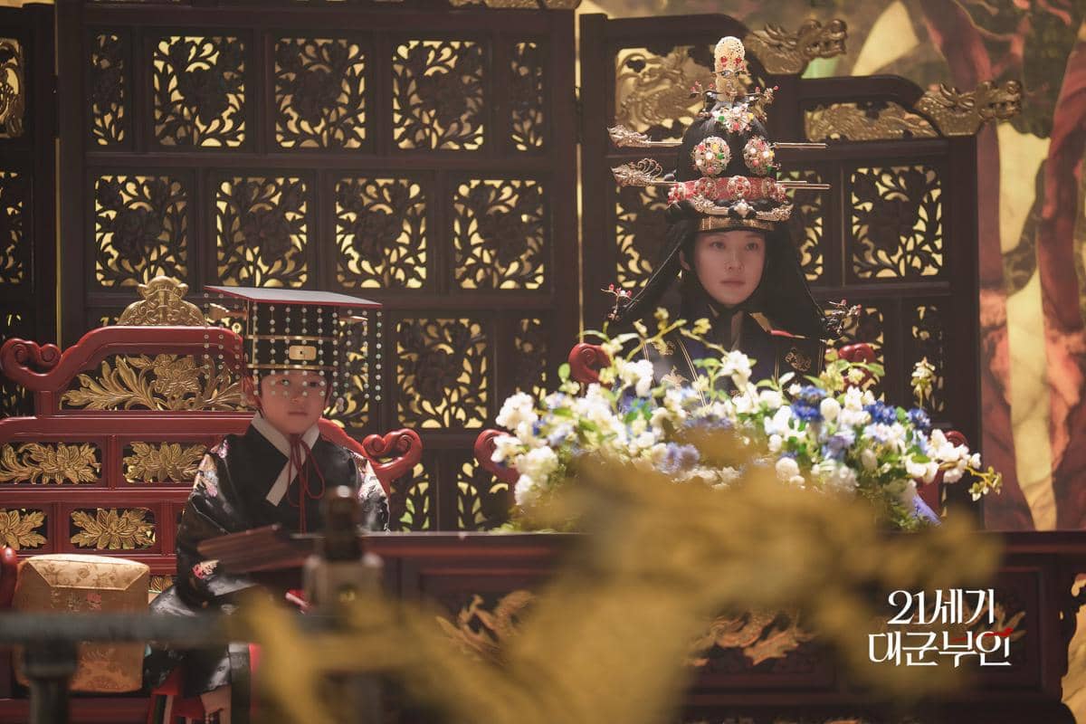 Dua karakter mengenakan pakaian kerajaan tradisional Korea duduk di singgasana berhias ukiran emas dalam adegan drama Perfect Crown.