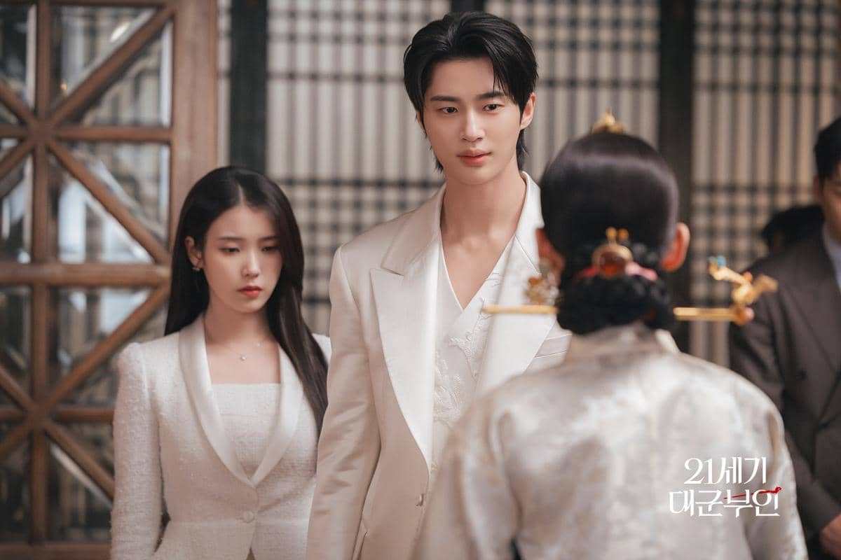 Dua karakter berpakaian formal putih berdiri di depan seorang wanita berbusana tradisional dalam adegan drama Perfect Crown.