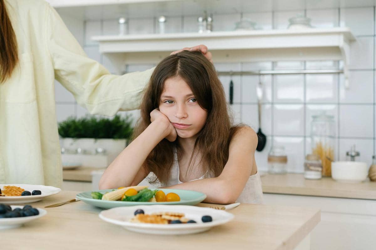 Seorang anak perempuan duduk di meja makan dengan ekspresi murung di depan piring berisi sayuran dan buah di dapur rumah.