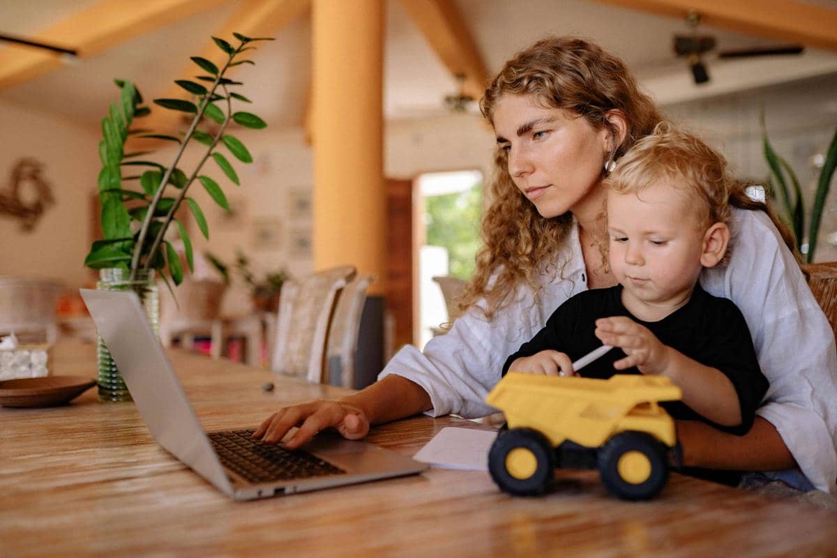 Seorang perempuan bekerja di laptop sambil mendampingi anak kecil yang bermain dengan mainan truk kuning di meja kayu.