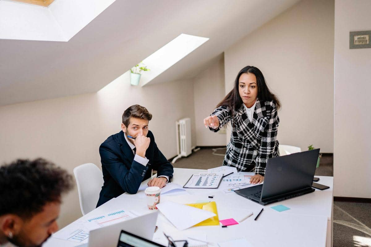 Seorang wanita menunjuk dengan ekspresi tegas kepada rekan kerja pria di ruang kantor, dengan dokumen dan laptop di atas meja.