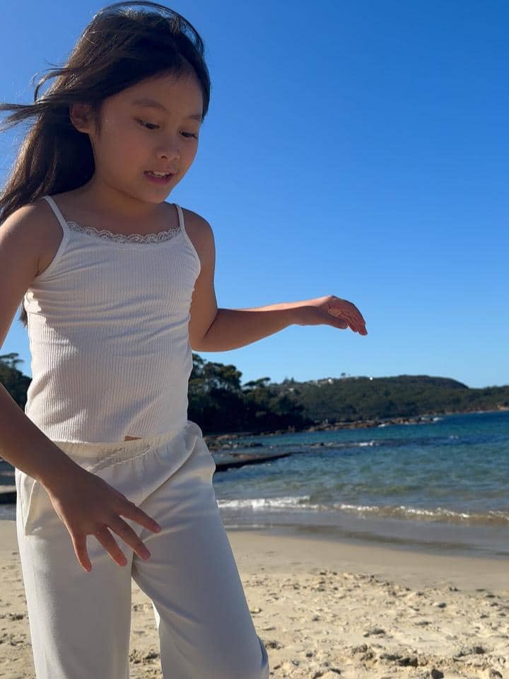 Seorang anak perempuan mengenakan pakaian putih bermain di tepi Pantai Balmoral dengan latar laut biru dan langit cerah.