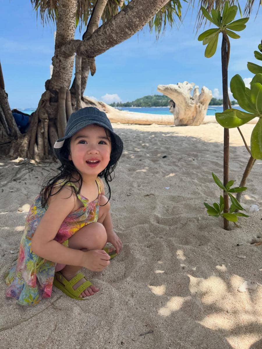 Seorang anak kecil mengenakan topi abu-abu dan gaun warna-warni bermain di pasir pantai berlatar laut biru di Lombok.