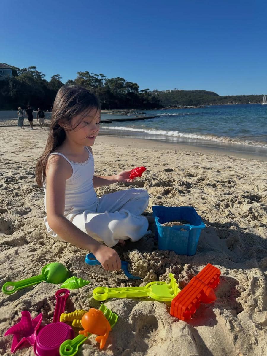 Seorang anak bermain pasir dengan mainan berwarna cerah di tepi Pantai Balmoral di bawah langit biru cerah.