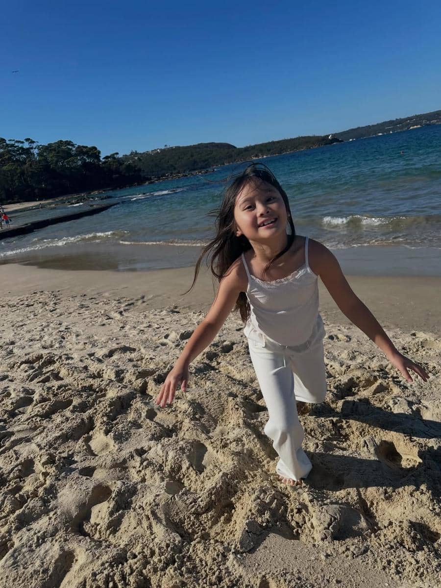 Seorang anak perempuan berlari di pasir Pantai Balmoral dengan latar laut biru dan langit cerah di siang hari.