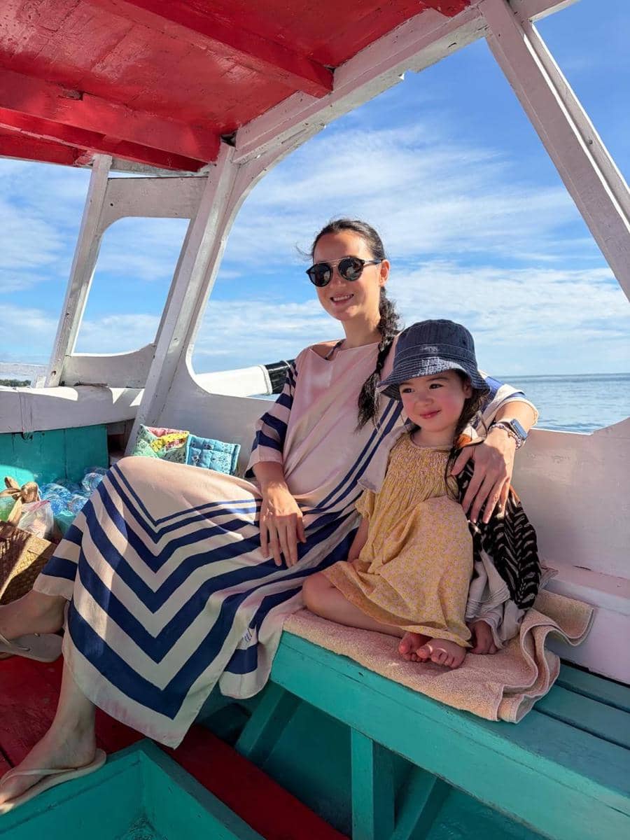Seorang wanita mengenakan gaun bermotif garis duduk di perahu bersama anak kecil bergaun kuning dan topi, dengan latar laut biru di Lombok.