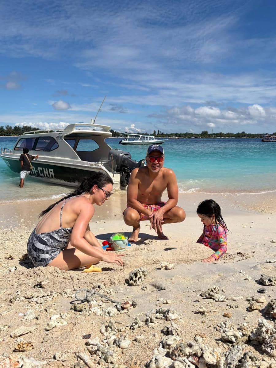 Seorang wanita, pria, dan anak kecil bermain pasir di tepi pantai berpasir putih dengan latar laut biru dan perahu di Lombok.