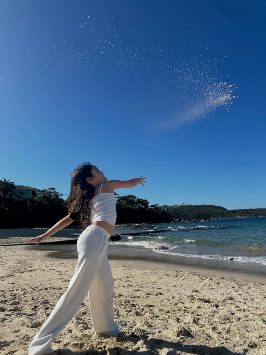 Seorang perempuan berpakaian putih melempar pasir ke udara di tepi Pantai Balmoral dengan latar langit biru cerah dan ombak tenang.