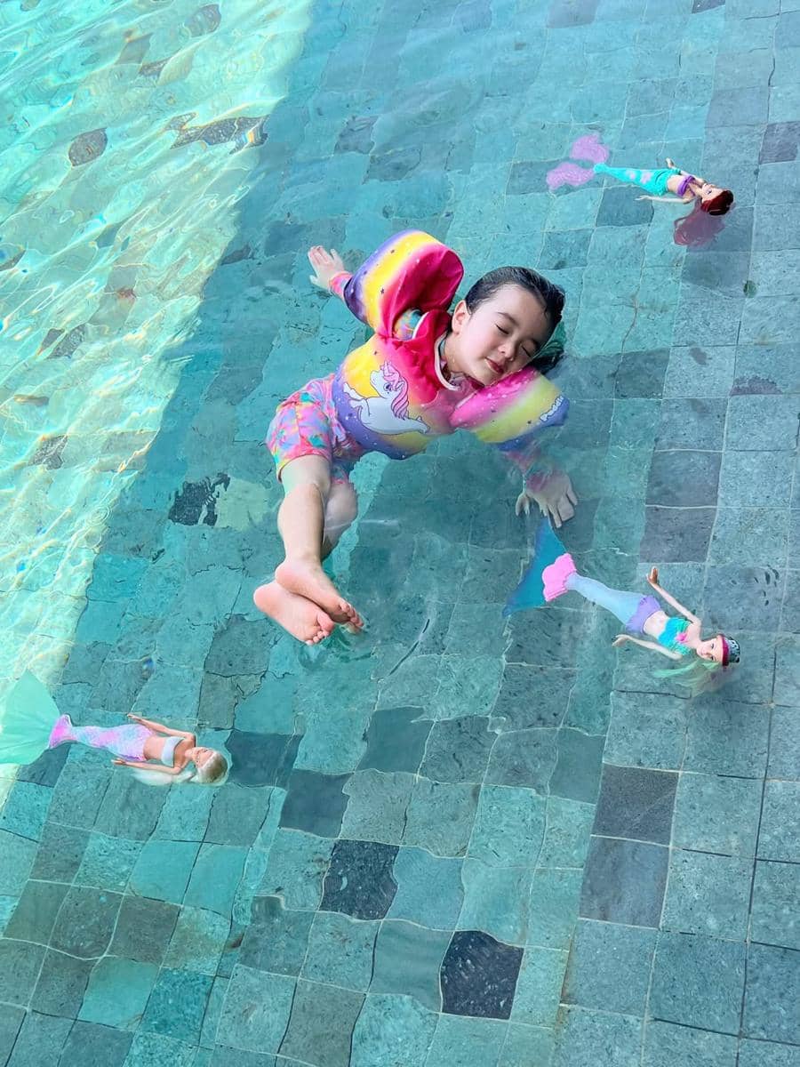 Seorang anak kecil mengenakan pelampung warna-warni sedang berenang santai di kolam renang bersama beberapa boneka putri duyung.