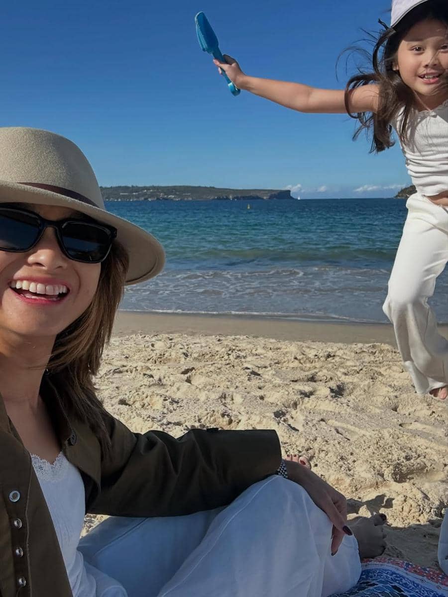 Seorang wanita mengenakan topi dan kacamata hitam tersenyum di pantai berpasir, sementara seorang anak bermain riang di dekatnya dengan latar laut biru.