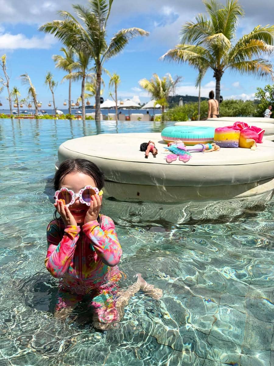 Seorang anak mengenakan baju renang warna-warni dan kacamata berbentuk hati bermain di kolam renang dengan latar pohon kelapa di Lombok.
