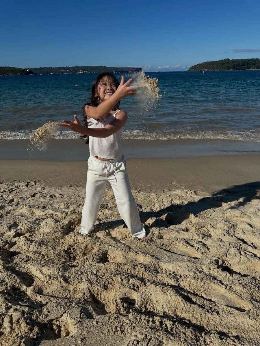 Seorang anak perempuan bermain pasir dengan gembira di tepi Pantai Balmoral berlatar laut biru dan langit cerah.