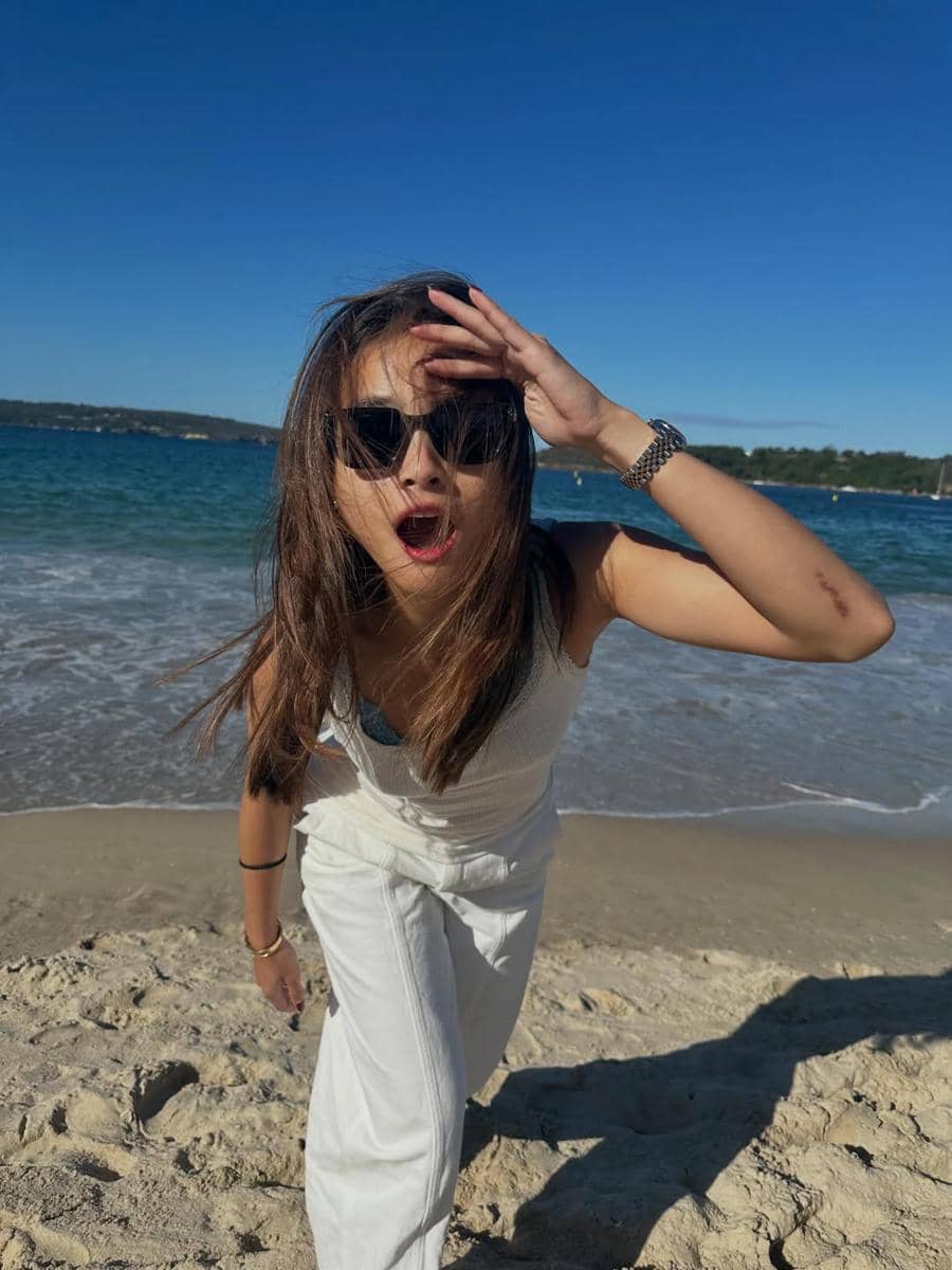 Seorang perempuan mengenakan kacamata hitam dan pakaian putih berdiri di tepi pantai berpasir dengan latar laut biru dan langit cerah.