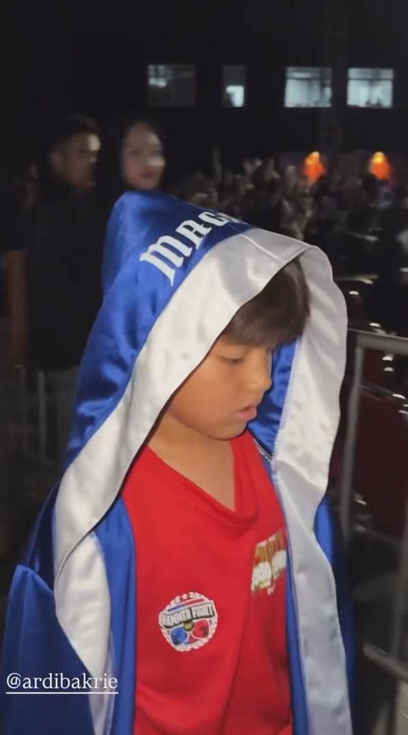 Seorang anak mengenakan jubah tinju biru dengan tudung putih dan kaus merah, berjalan di area pertandingan tinju dengan penonton di latar belakang.