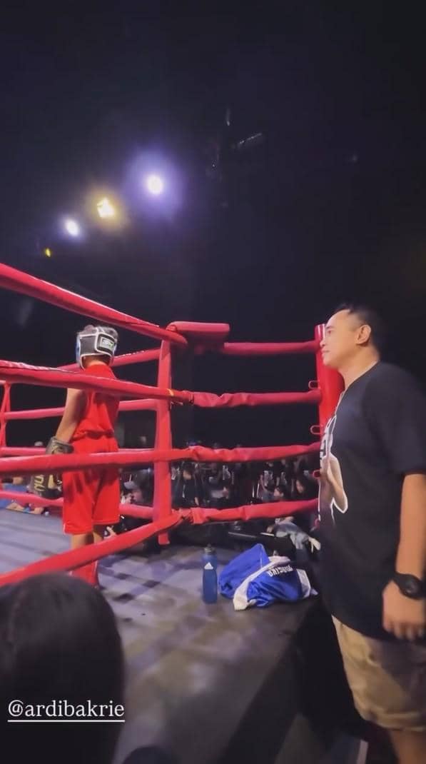 Seorang anak mengenakan perlengkapan tinju berwarna merah berdiri di sudut ring dengan pelatih di sisinya di arena pertandingan.