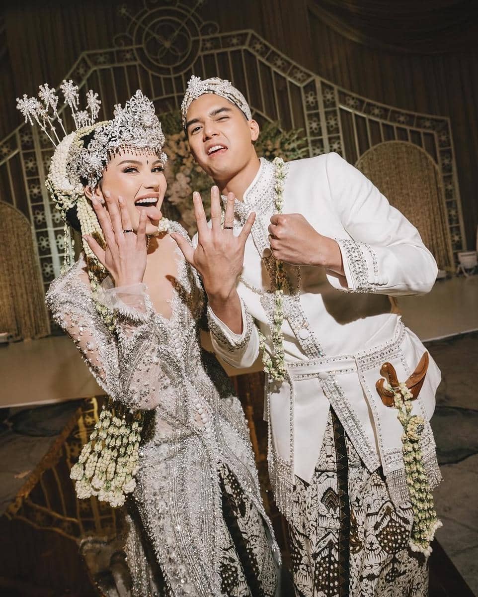 Pasangan pengantin mengenakan busana adat Jawa modern berwarna putih dan perak dengan hiasan kepala serta rangkaian melati di hari pernikahan.