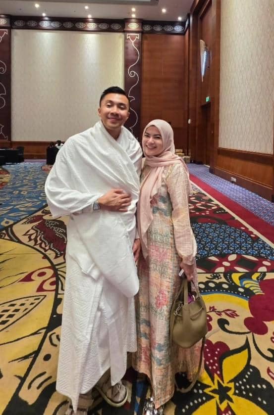 Seorang pria mengenakan pakaian ihram berdiri di samping seorang wanita berhijab dengan gaun panjang di dalam ruangan berkarpet motif warna-warni.