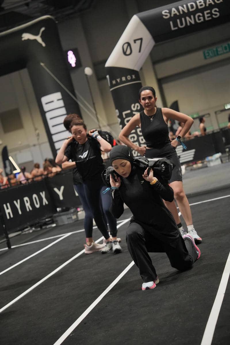 Beberapa peserta wanita melakukan latihan sandbag lunges di ajang Hyrox Hong Kong dengan suasana kompetitif di arena dalam ruangan.