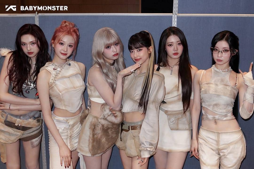 Enam anggota BABYMONSTER berpose bersama mengenakan busana bernuansa krem dan beige di depan latar biru abu-abu.