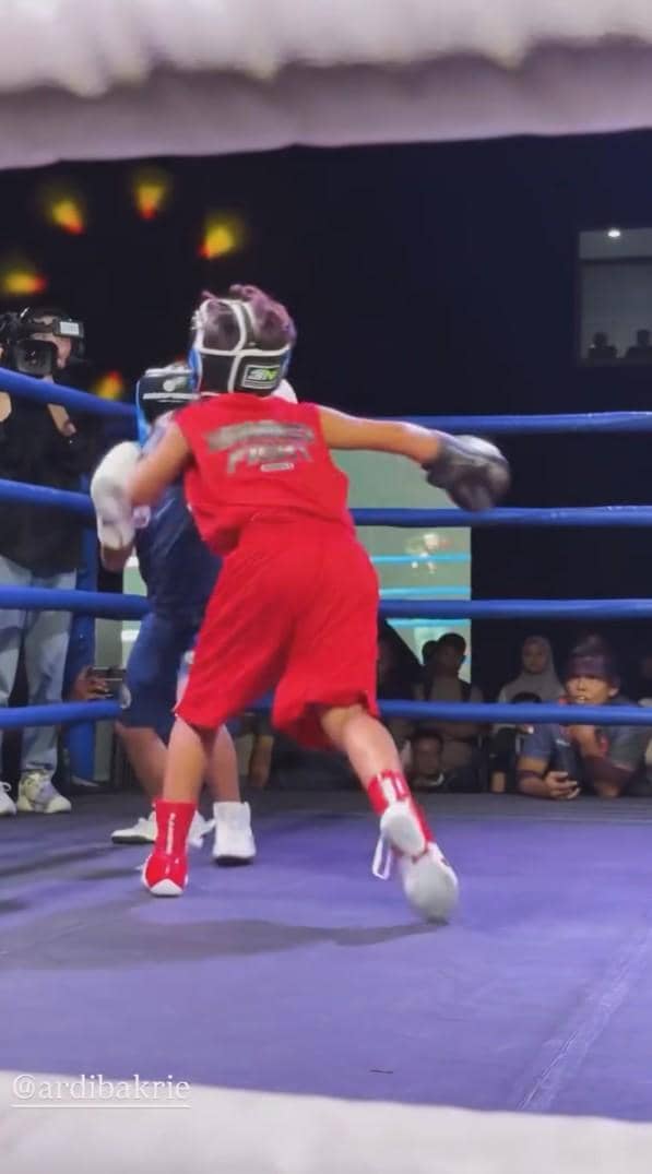 Seorang anak mengenakan perlengkapan tinju berwarna merah sedang bertanding di atas ring melawan lawan berpakaian biru di depan penonton.