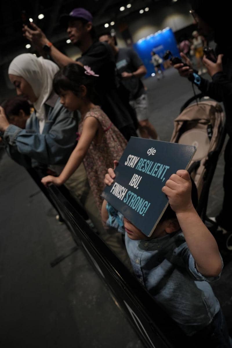 Seorang anak memegang papan bertuliskan semangat untuk ibunya di acara Hyrox Hong Kong, dikelilingi keluarga dan penonton lain.