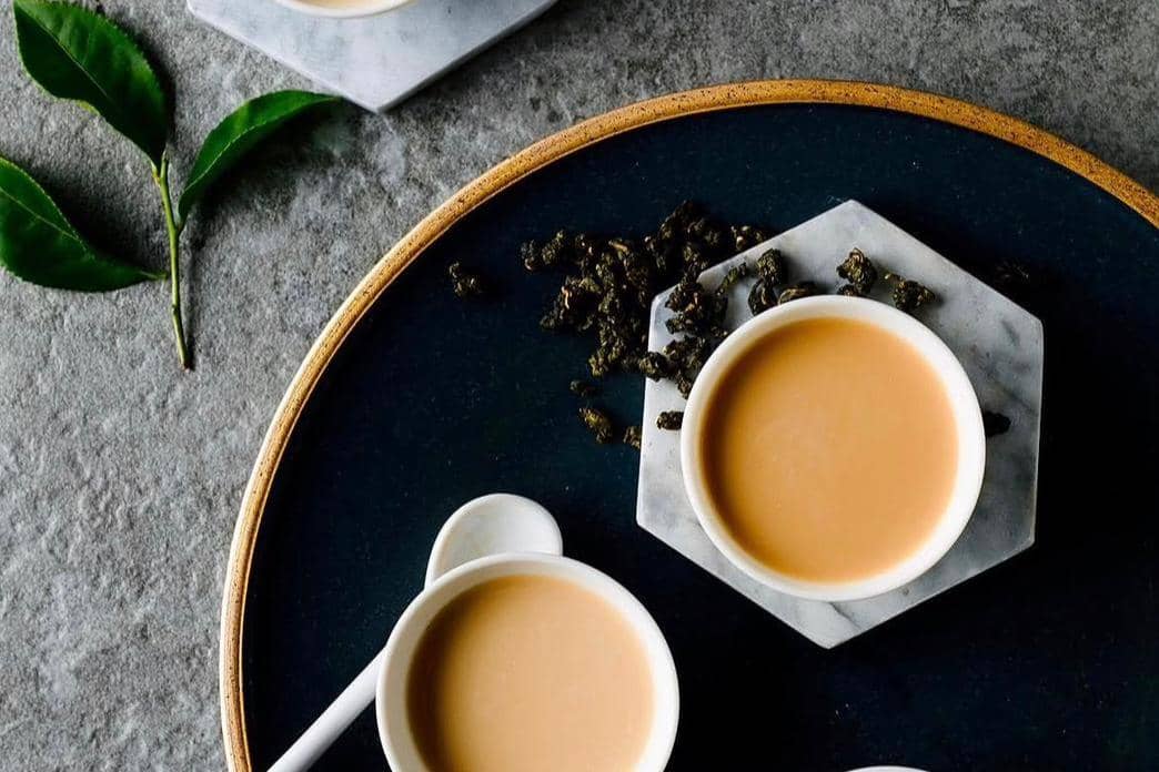 Dua cangkir teh susu disajikan di atas tatakan marmer dengan daun teh kering dan daun teh segar di permukaan abu-abu.