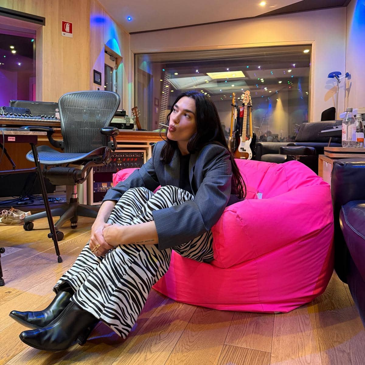 Seorang perempuan duduk di bean bag berwarna pink di studio musik dengan peralatan rekaman dan gitar di latar belakang.