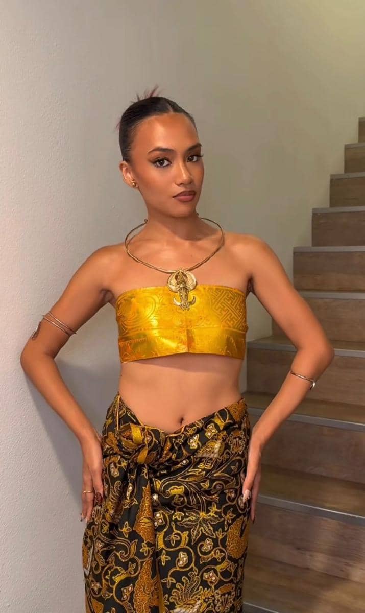 Seorang wanita mengenakan atasan kain emas dan kain batik hitam emas berdiri di depan tangga dengan gaya elegan.