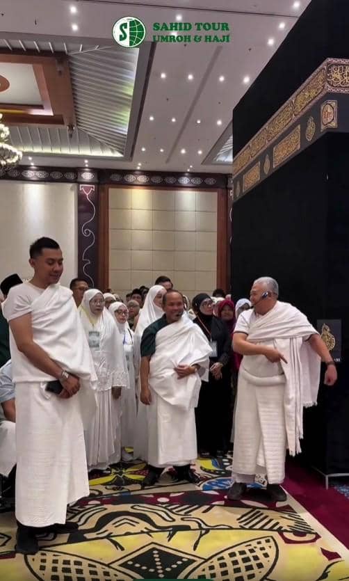 Sekelompok calon jamaah haji mengenakan pakaian ihram sedang mengikuti bimbingan manasik haji di dalam ruangan dengan replika Kakbah.