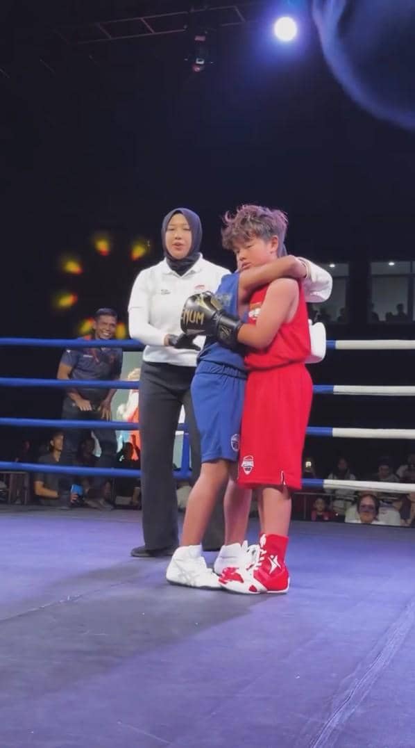 Dua anak mengenakan perlengkapan tinju saling berpelukan di atas ring dengan wasit berdiri di dekat mereka di bawah sorotan lampu.