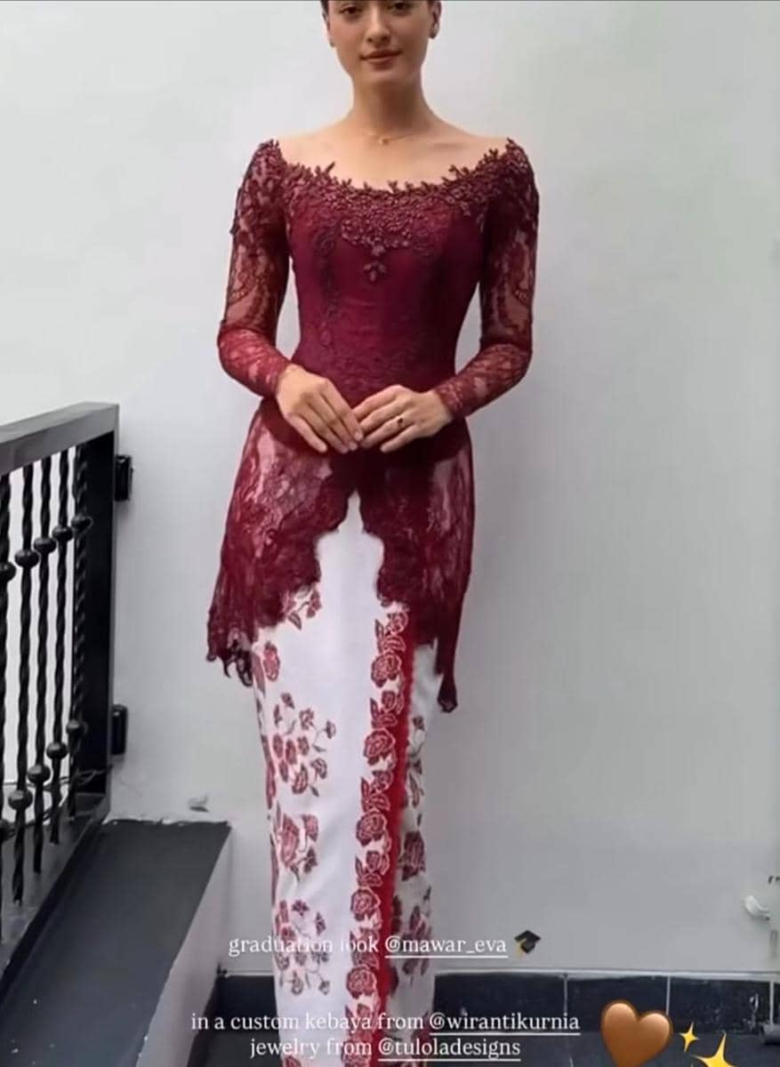 Seorang perempuan mengenakan kebaya merah marun dengan rok bermotif bunga putih merah, berdiri di dekat dinding putih saat wisuda.