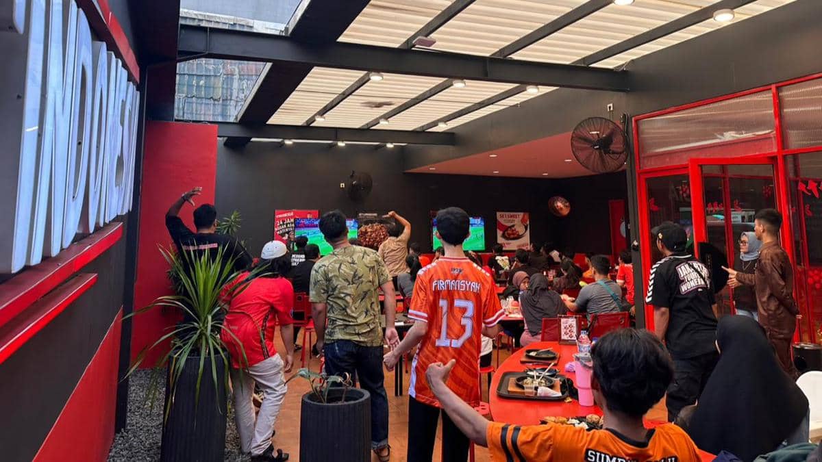 Suasana nobar pertandingan Persija melawan Persib di sebuah kafe dengan penggemar mengenakan jersey oranye dan hitam.