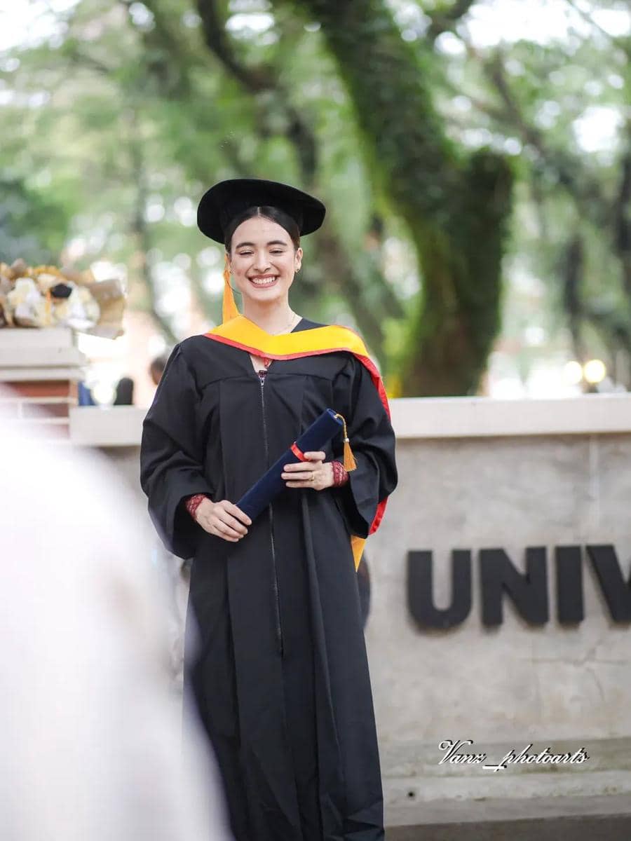 Seorang wisudawati mengenakan toga hitam dengan selempang kuning dan merah, tersenyum sambil memegang ijazah di area kampus.
