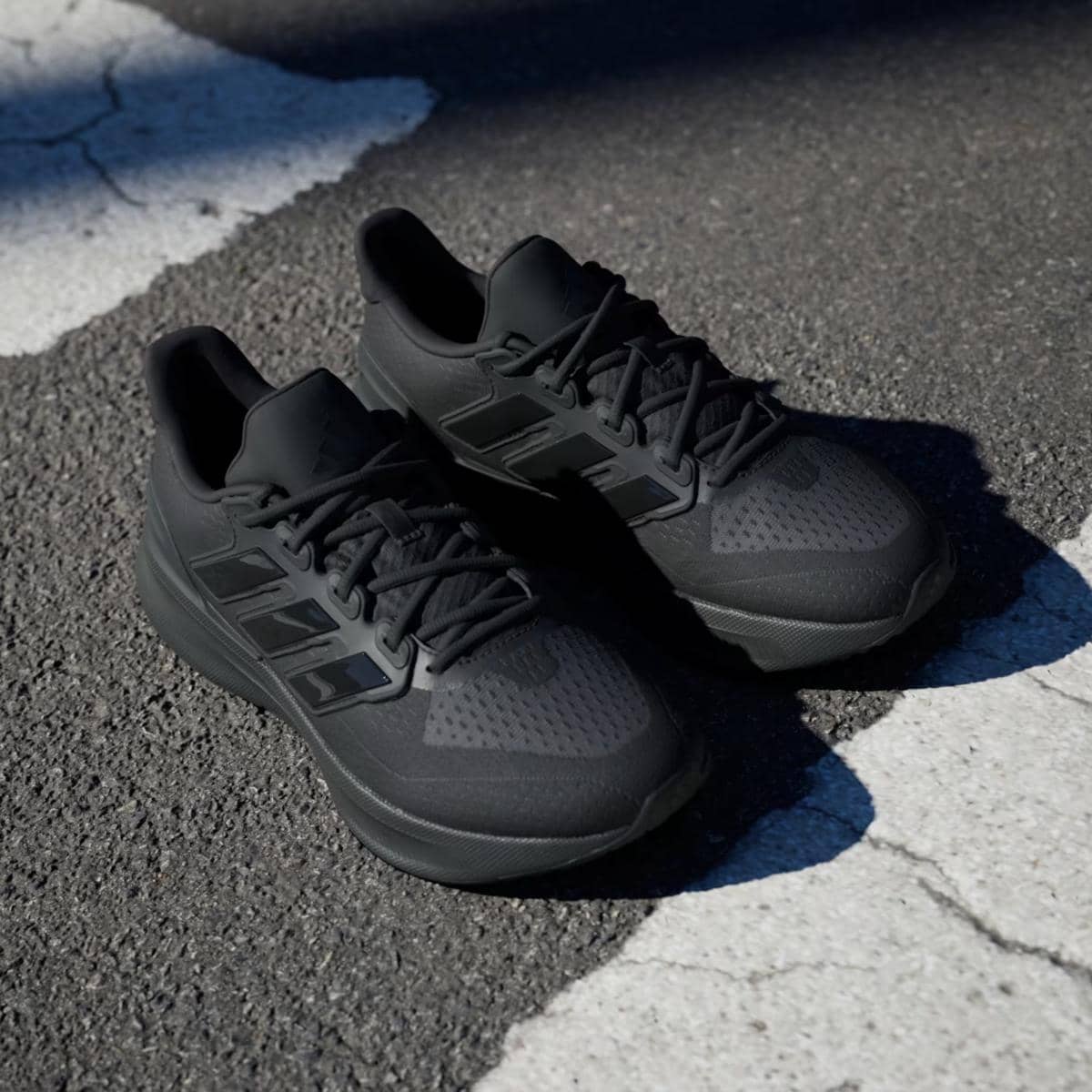 Sepasang sepatu lari Adidas Ultrarun 5 warna hitam pekat diletakkan di atas permukaan jalan beraspal dengan cahaya matahari sebagian.