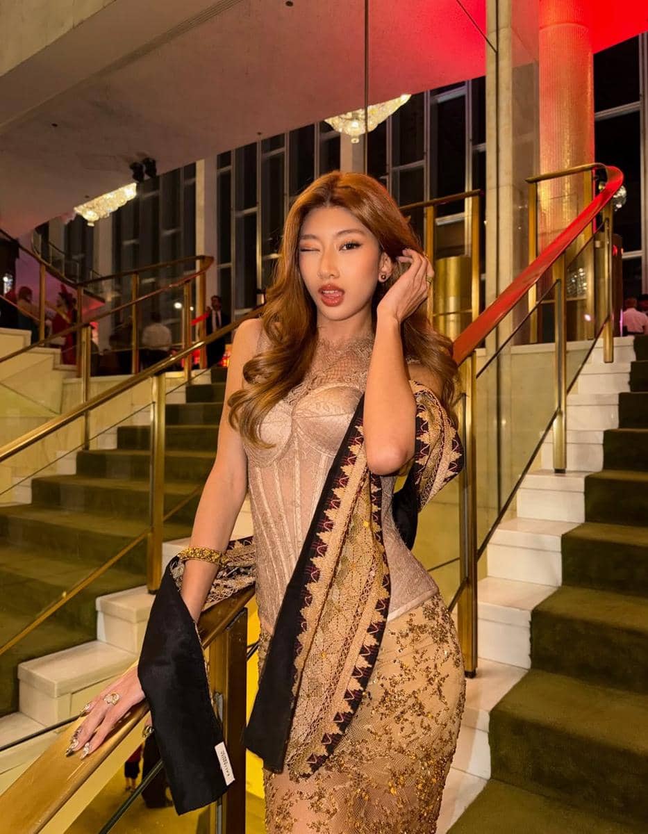 Seorang perempuan berpose di tangga dengan gaun berwarna emas dan detail bordir, di dalam gedung dengan pencahayaan hangat.