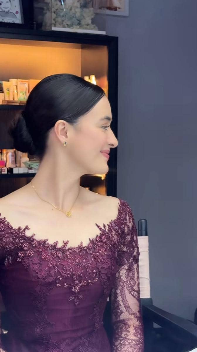Seorang perempuan mengenakan kebaya ungu marun berhias renda dengan rambut disanggul rapi, tersenyum sambil menoleh ke samping.