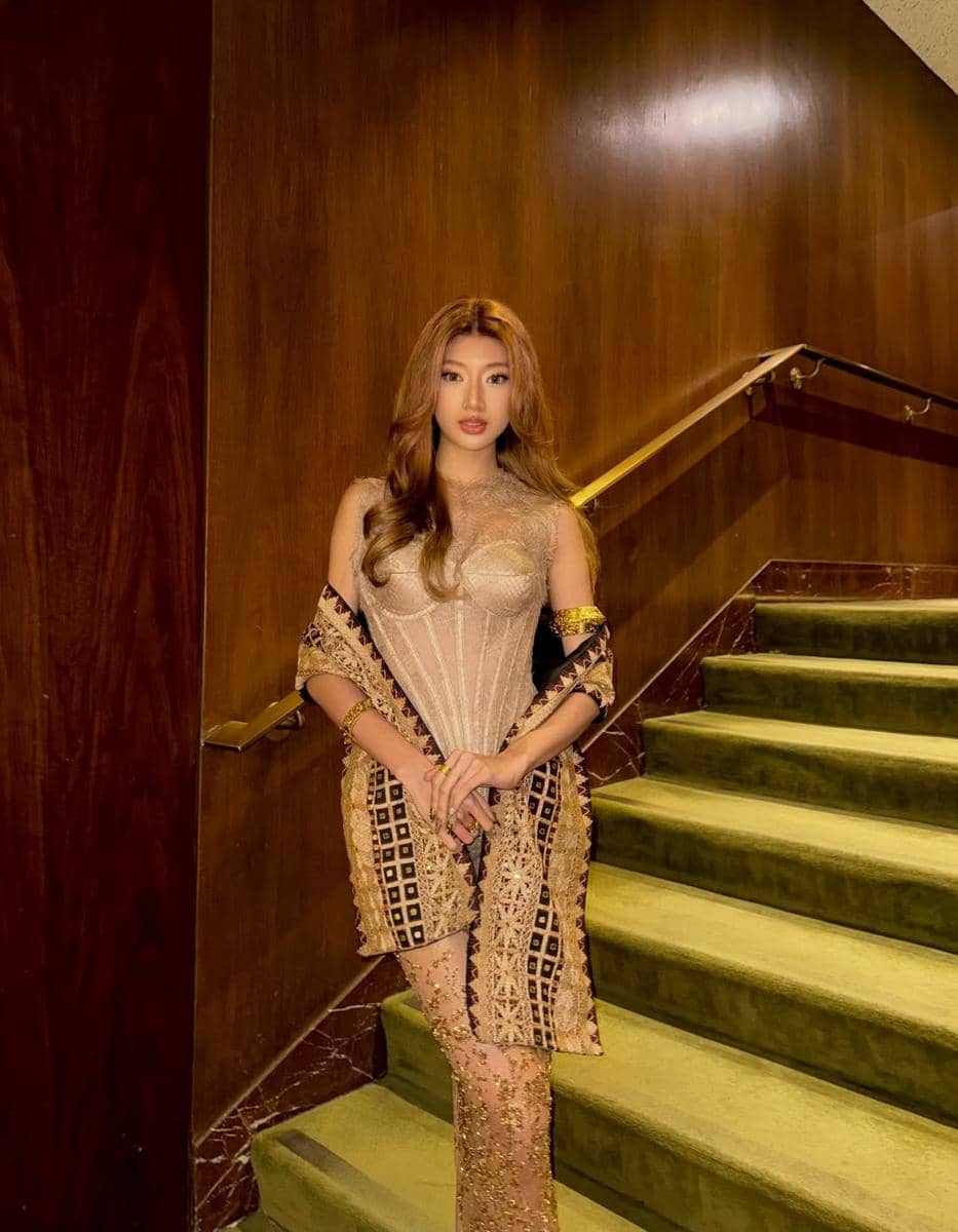 Seorang wanita berpose di tangga berkarpet hijau dengan dinding kayu, mengenakan gaun berwarna emas dan selendang bermotif batik.